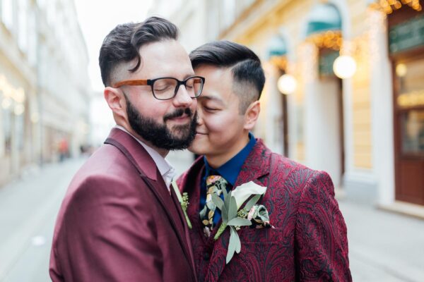 Zwei Männer in bordeauxroten Anzügen lehnen in einer romantischen Pose aneinander