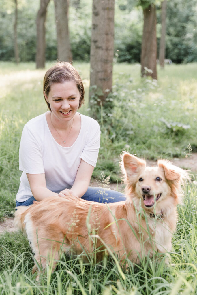 Frau im weißen Shirt hockt neben einem hellbraunen Hund auf einer Wiese.
